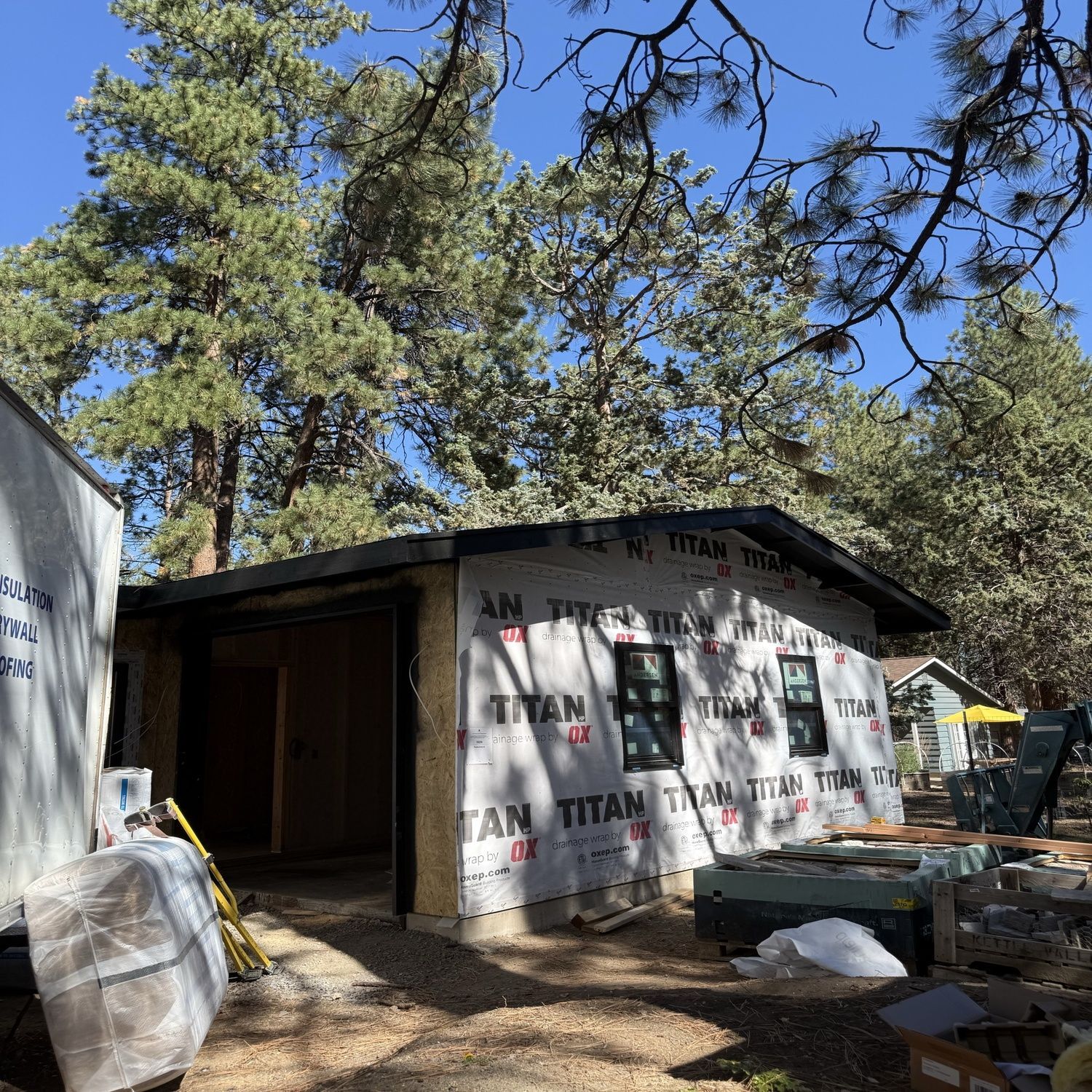 Partially built shed wrapped in insulation under tall trees, with construction materials scattered around.