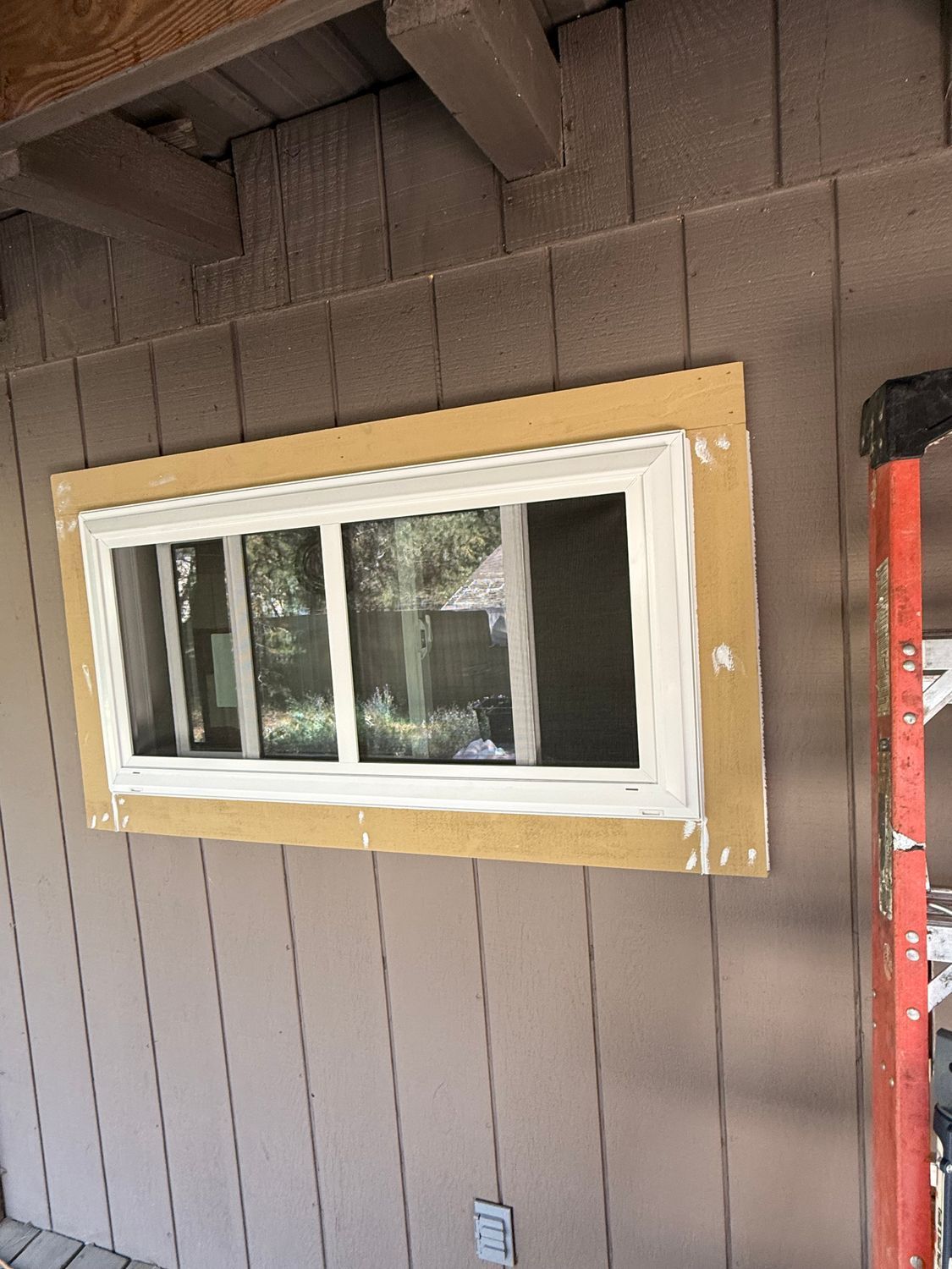 Small window set in brown vertical siding, framed with unfinished wood trim on a porch wall.