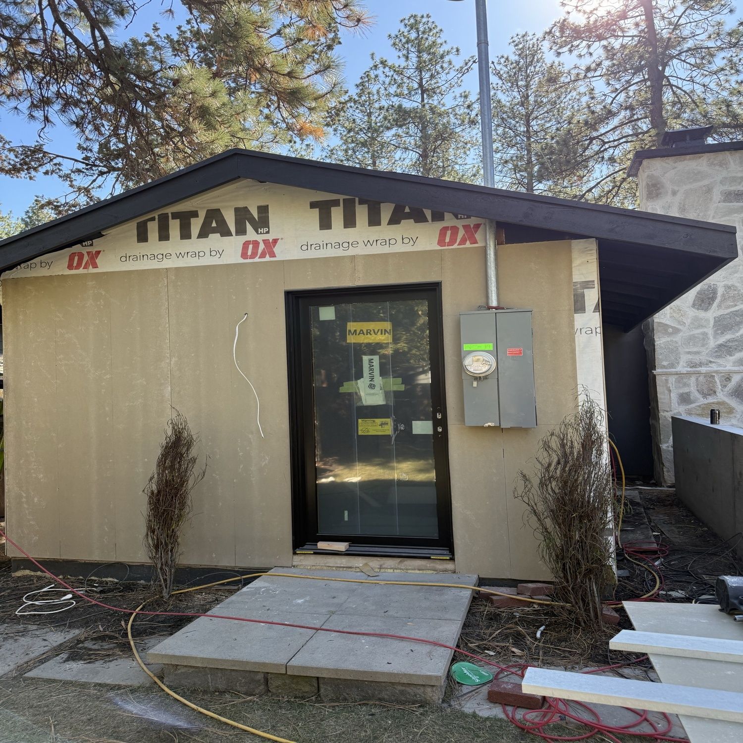 Small beige building with a black door, red roof trim, and a concrete walkway in a wooded area.