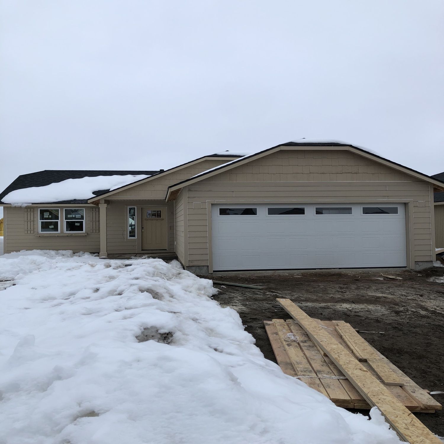 Snow-covered beige suburban house with a two-car garage and a driveway under construction.