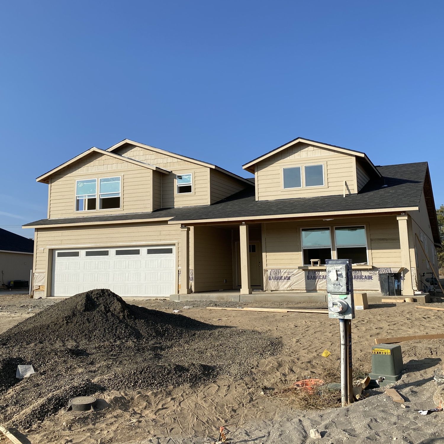 New beige two-story house under construction with garage, dirt front yard, and clear blue sky