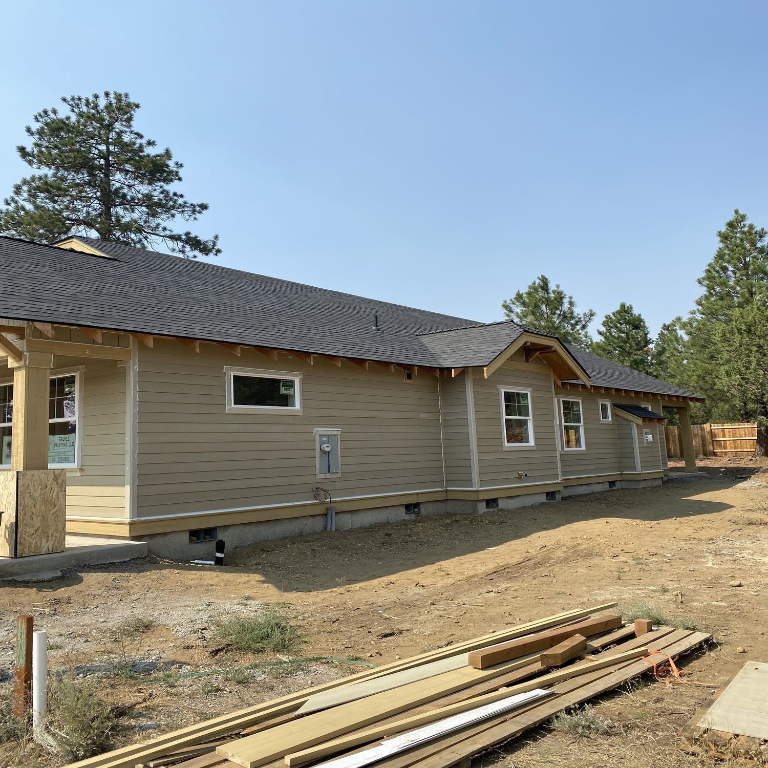 Single-story house under construction with tan siding, dark roof, and lumber stacked in the foreground.