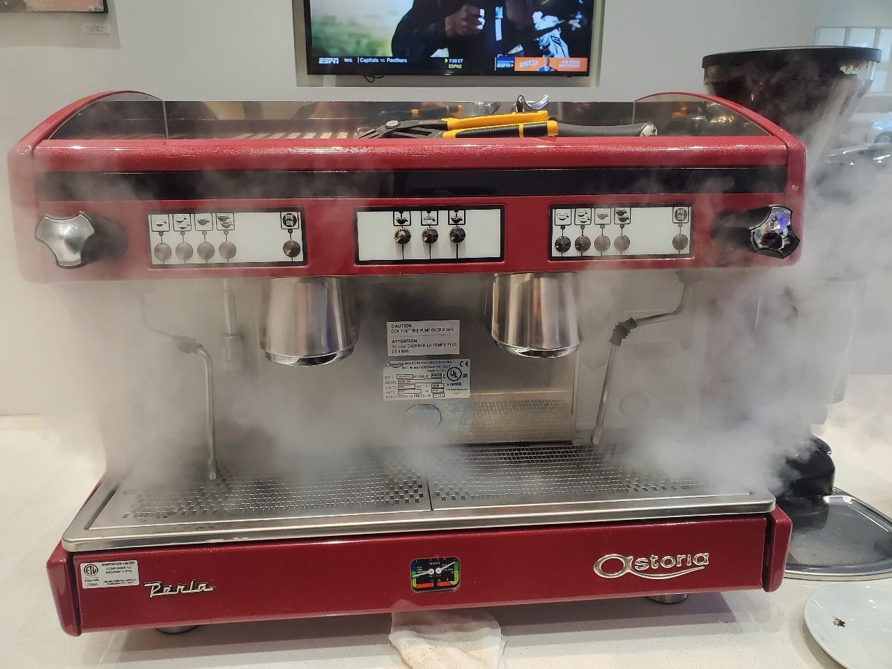 Red espresso machine steaming hot water into a cup, with a TV screen above it.