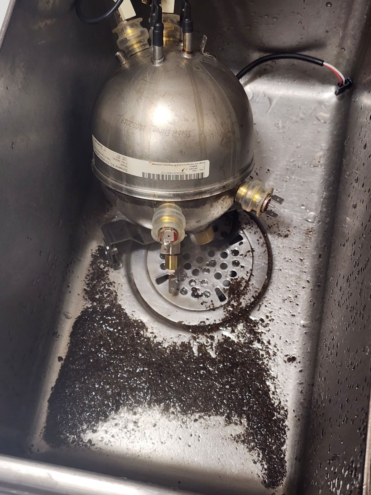 Metal tank with fittings in a stainless-steel sink, surrounded by spilled dark granules.