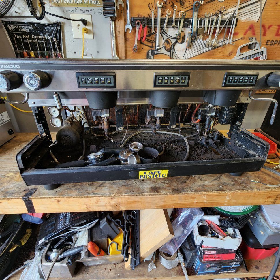Dusty espresso machine on a workbench in a cluttered workshop, with tools hanging on the wall behind it.
