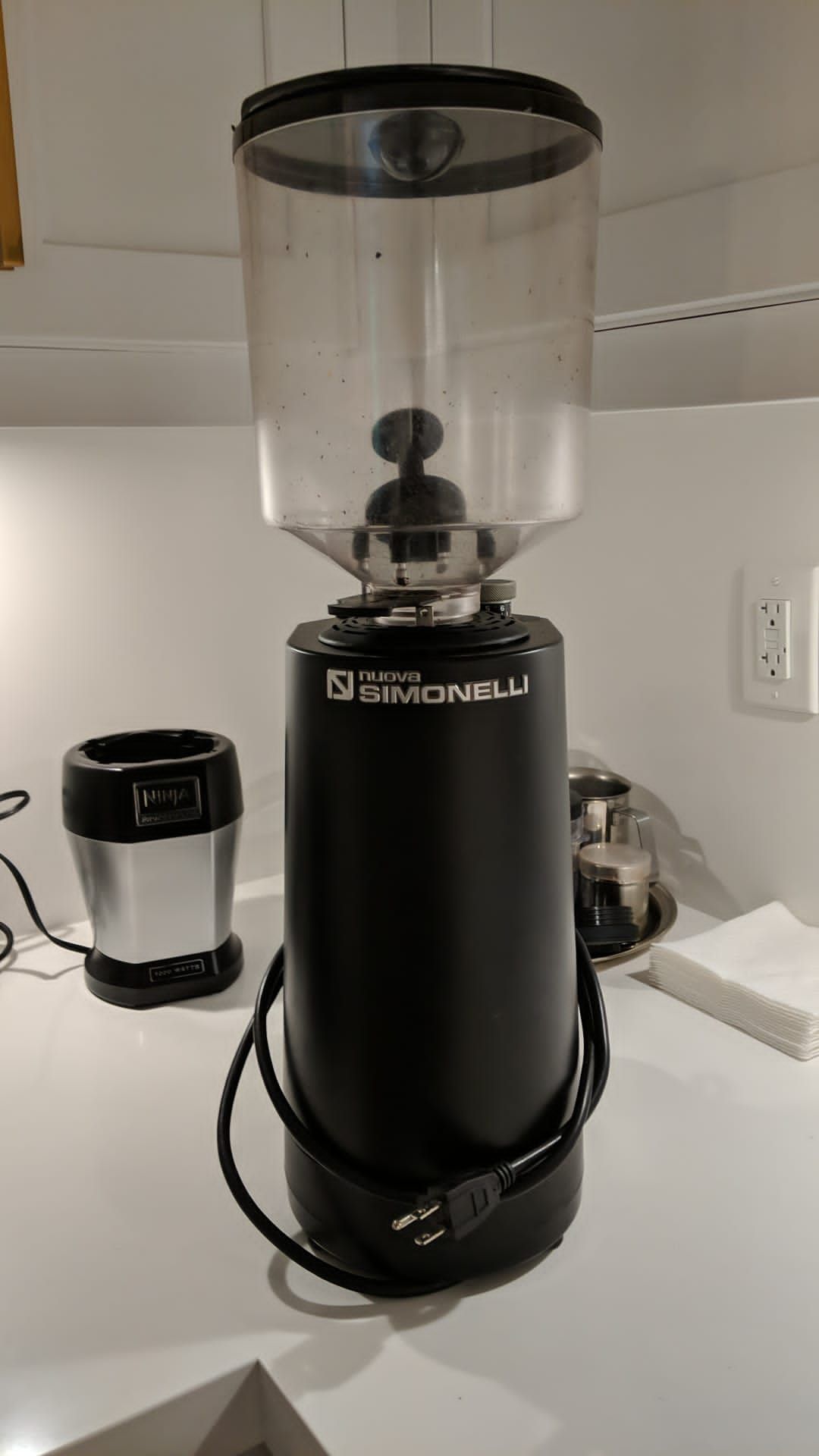 Black coffee grinder with clear bean hopper on a kitchen counter, next to a small appliance and outlet.