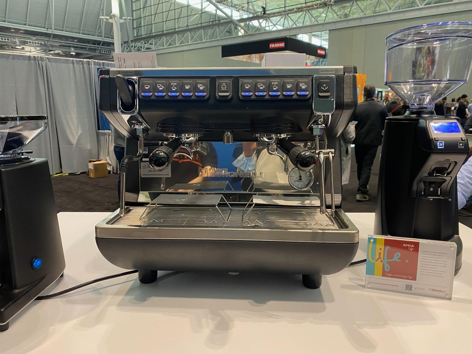 Stainless espresso machine on a counter at a coffee expo, with display lights and cups nearby