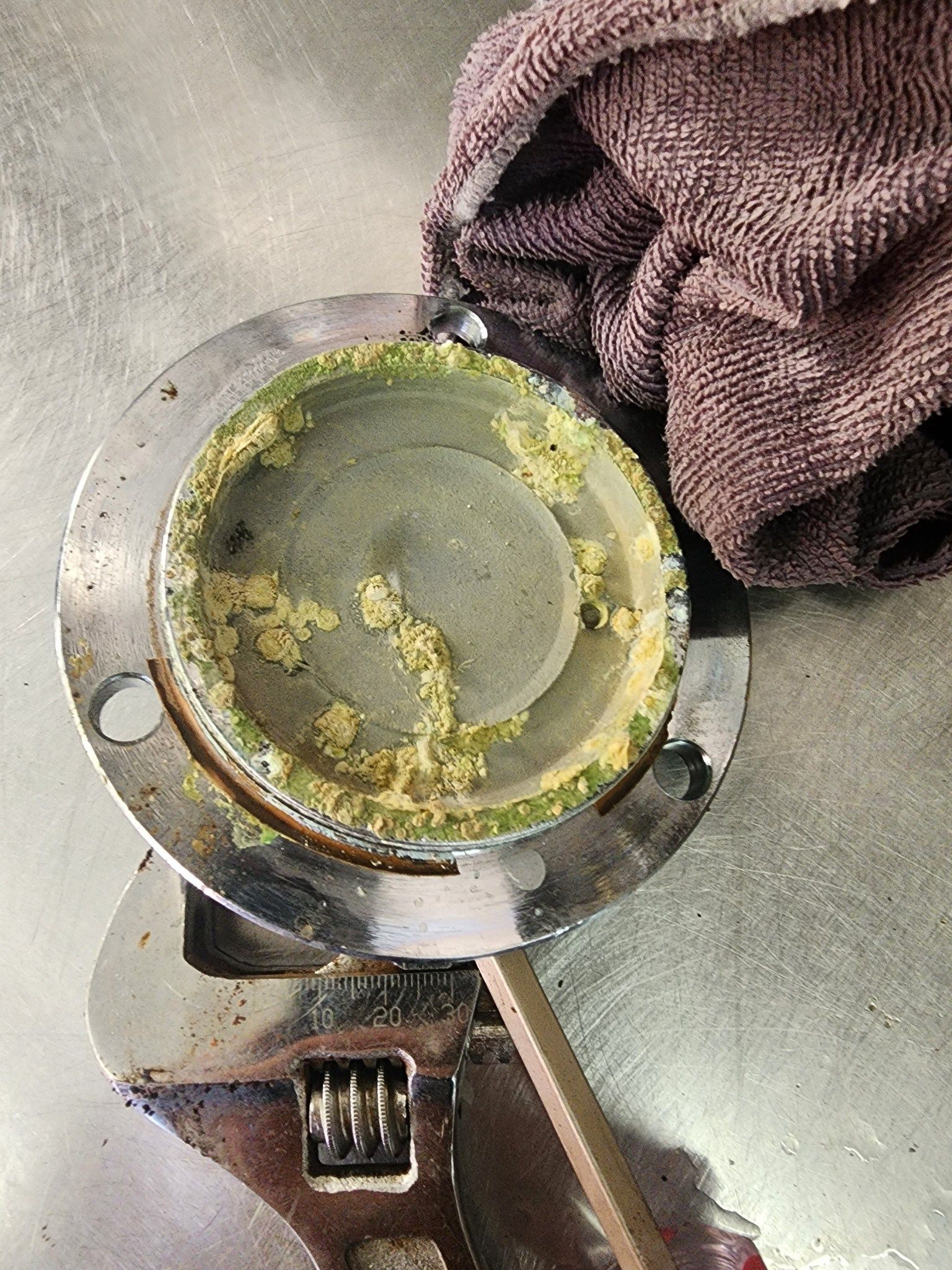 Dirty metal pot lid with green residue on a kitchen floor beside a purple towel