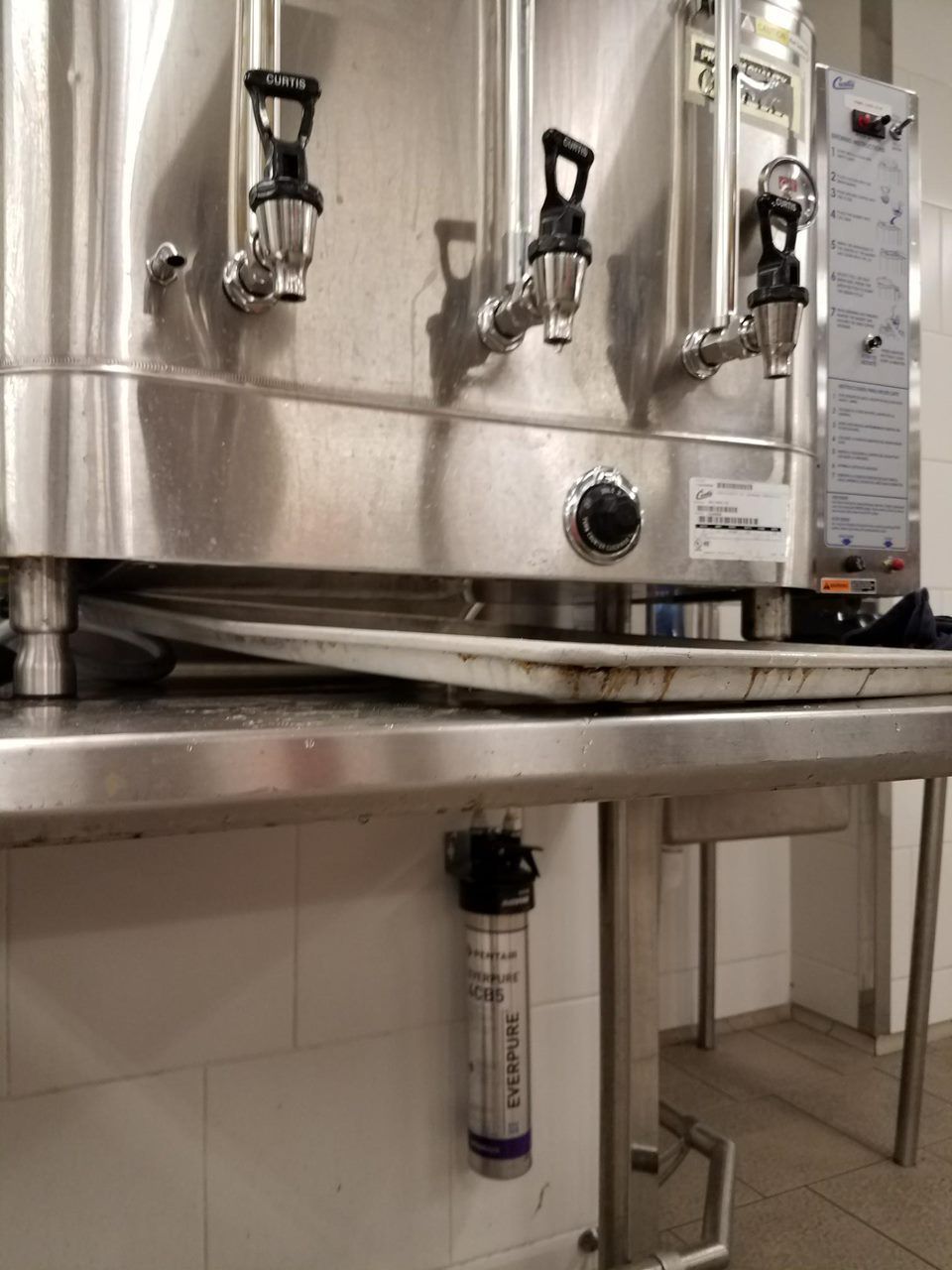 Stainless steel kitchen equipment with faucets and a pressure gauge in a tiled utility area
