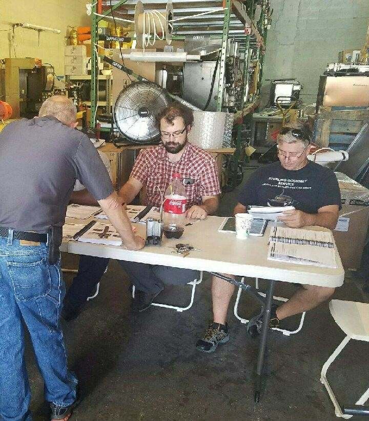 Three people seated at a table in a workshop, talking and writing amid metal equipment.