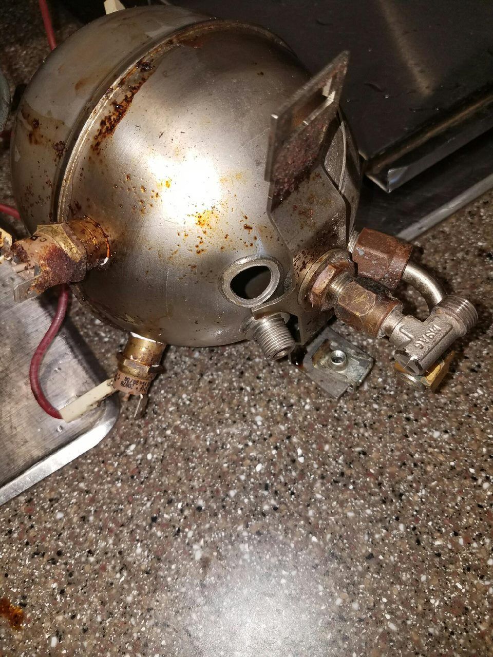 Metallic canister with rusted fittings lying on a wet, gravelly floor