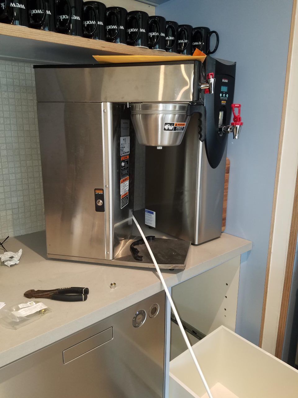 Coffee maker on a stainless counter in a kitchen, with a white hose leading into a bin below.