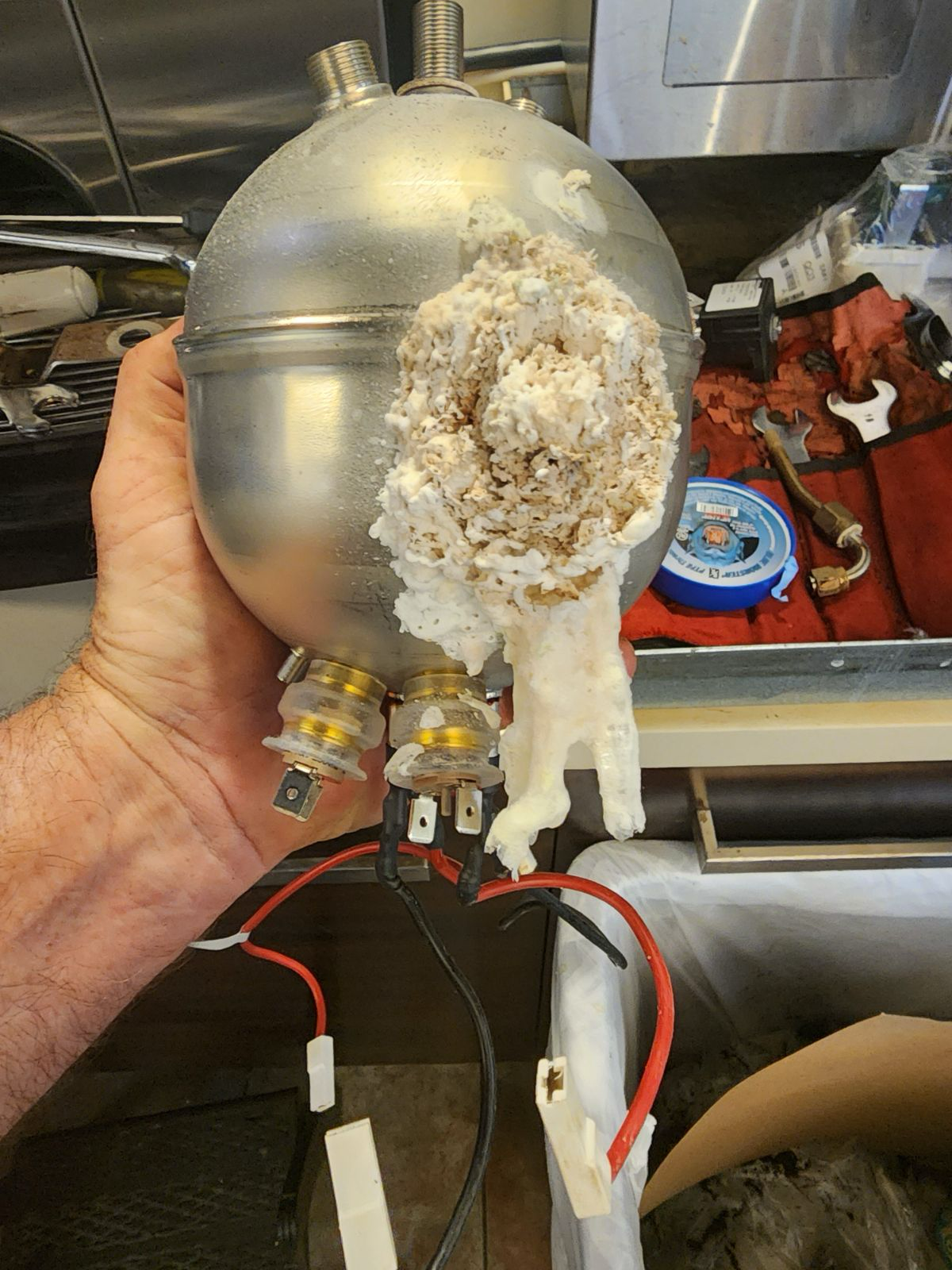 Hand holding a damaged silver metal device with a corroded section in a workshop setting