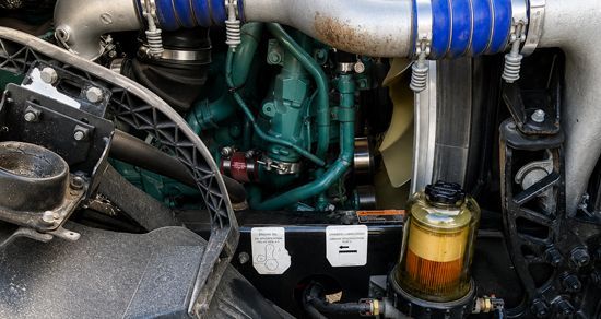 Close-up of a car engine bay with blue hoses, metal piping, and a clear fluid reservoir.