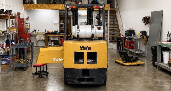 Yellow Yale forklift in a warehouse workshop, centered with metal shelves and equipment around it.