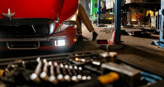 Red sports car in a garage with a mechanic working underneath, tools blurred in the foreground.