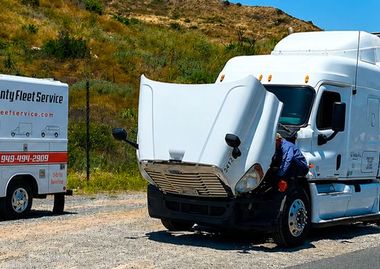 White semi truck with front cab cover lifted open on a roadside, with another service truck in the background.