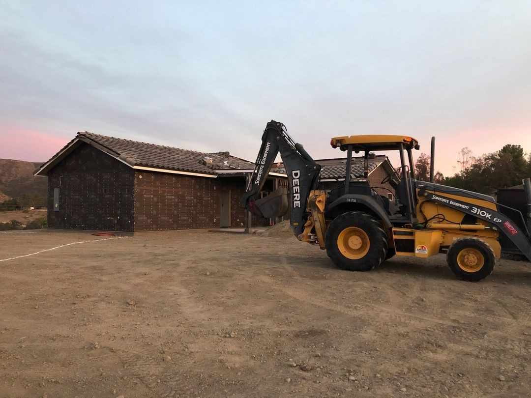 Yellow backhoe loader beside a small brick building at dusk on a dirt lot