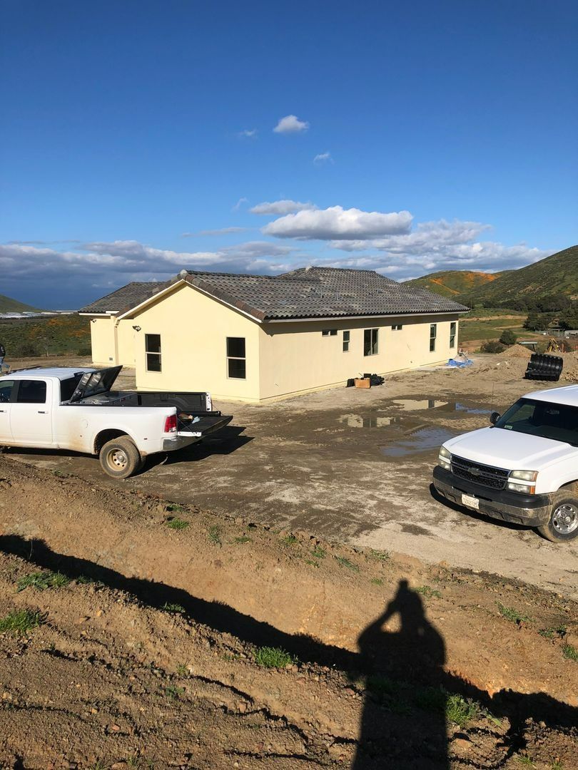 Yellow house under construction on a dirt lot, with two white pickup trucks and mountains in the background