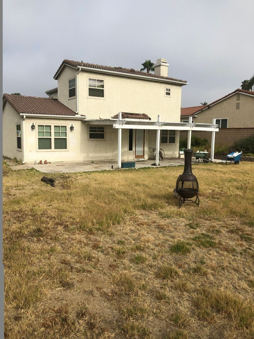 Suburban backyard with a two-story cream house, covered patio, and dry grassy lawn under cloudy skies