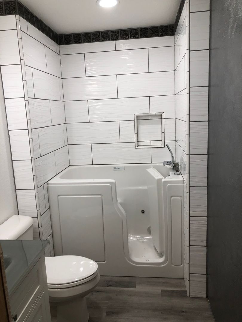 Small white tiled bathroom with a walk-in bathtub, toilet, and wall-mounted grab bar near a window.