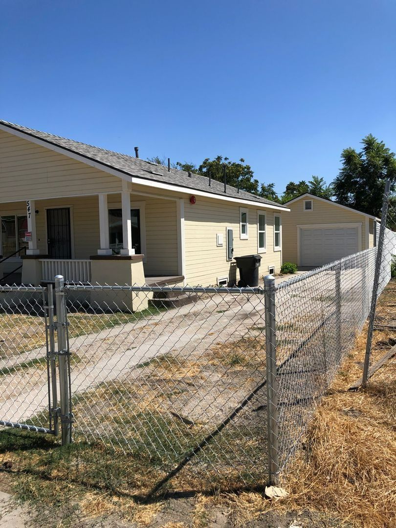 Damaged chain-link fence beside a beige house and detached garage on a dry, sloped yard