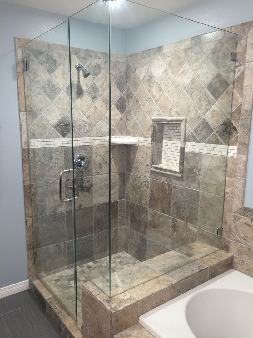 Glass walk-in shower with beige stone tile walls and chrome fixtures in a bathroom.