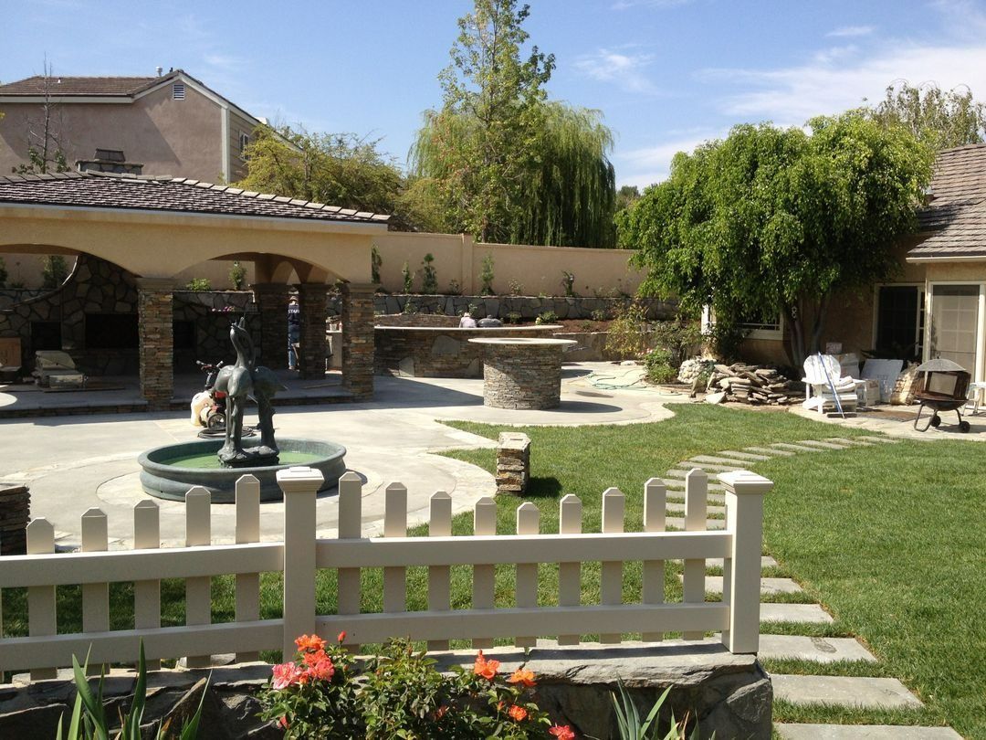 Courtyard with fountain, white picket fence, stone path, and lush green trees under a blue sky