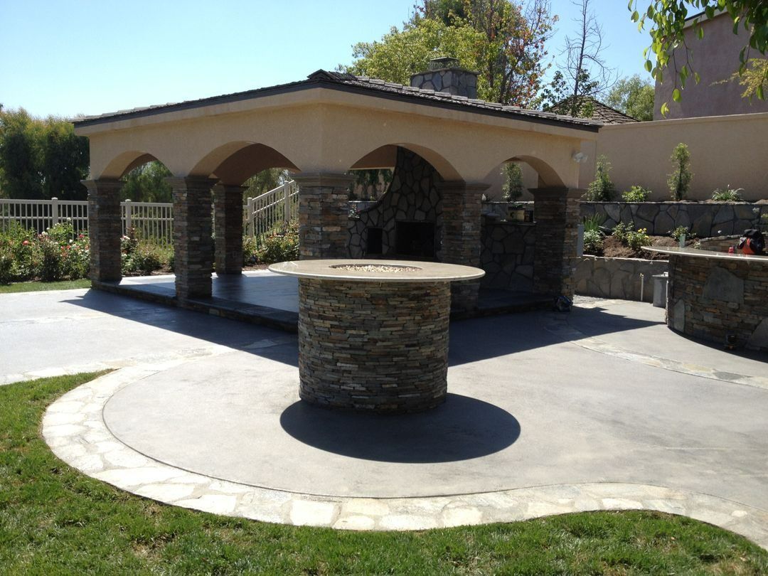 Outdoor stone pavilion with arched openings and a circular central table in a sunlit courtyard