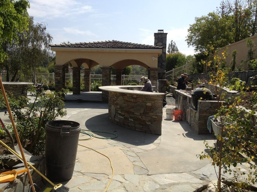 Outdoor stone patio with a circular stone fountain, arched pavilion, and people gathered in a sunny garden.