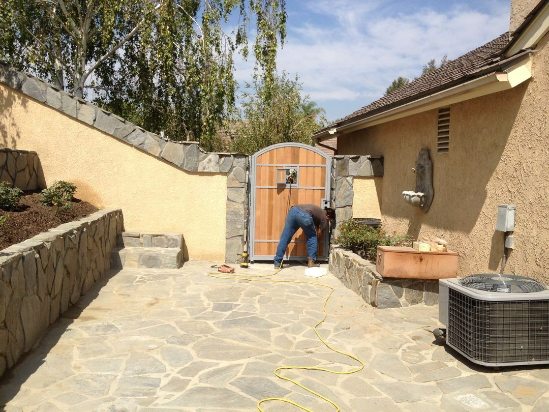 Person bending near an open gate in a sunlit courtyard with stone walls and paving.