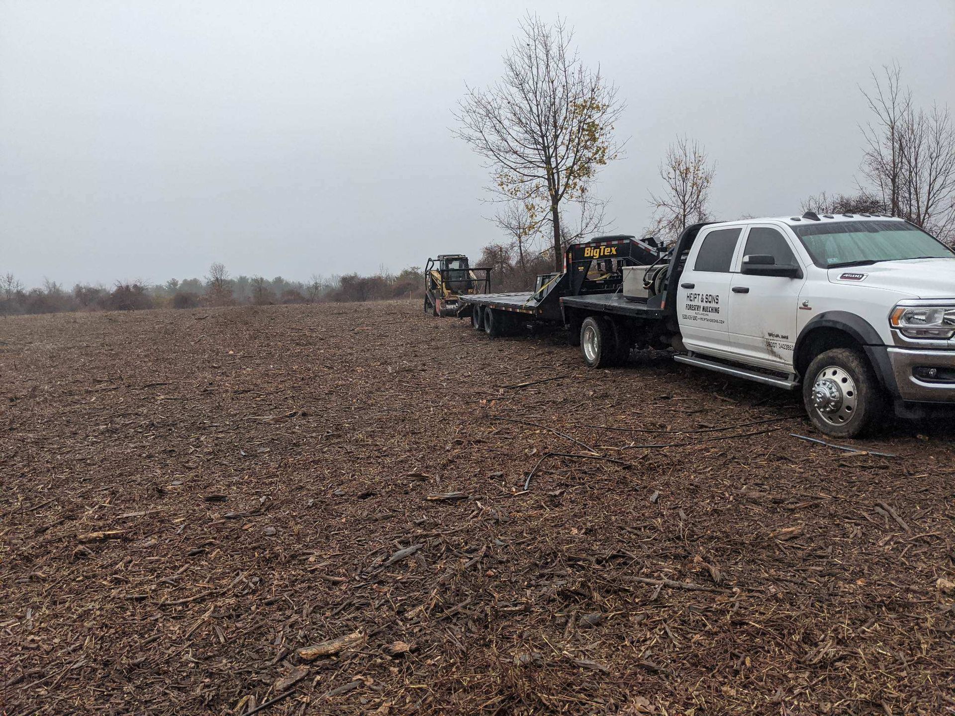 Excavator clearing a dirt road in a forest, piles of wood debris nearby, autumn foliage.