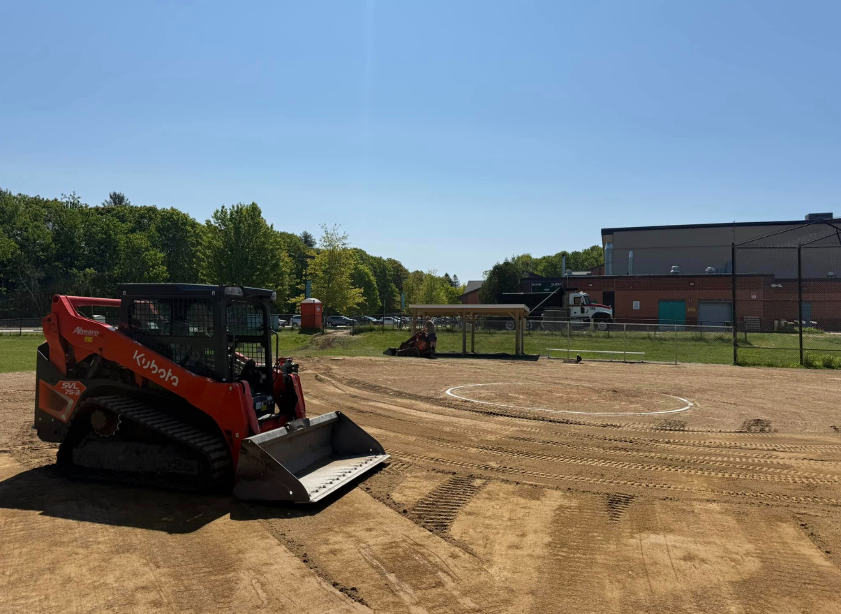 Red skid steer on a cleared construction site with a building and trees in the background under a blue sky.