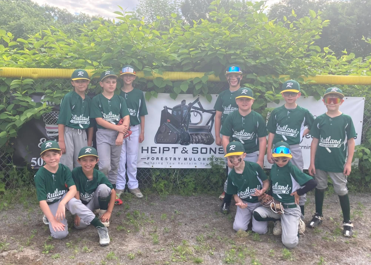 Baseball team in green jerseys and hats posing in front of a sign and bushes.