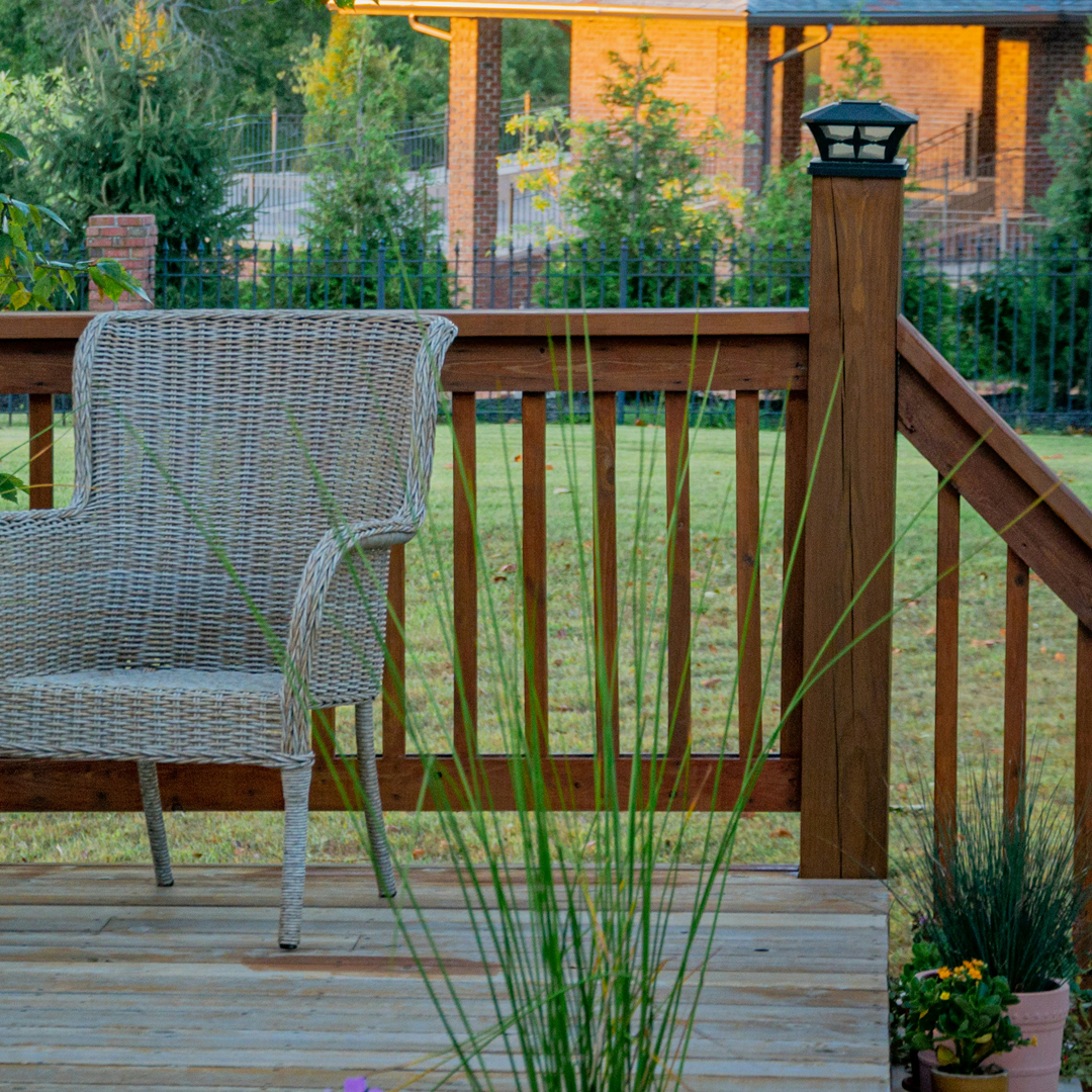 Wicker chair on a wooden deck with potted plants, railing, and a grassy yard beyond.