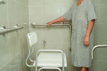 Person in hospital gown standing beside a shower chair and grab bars in a tiled bathroom.