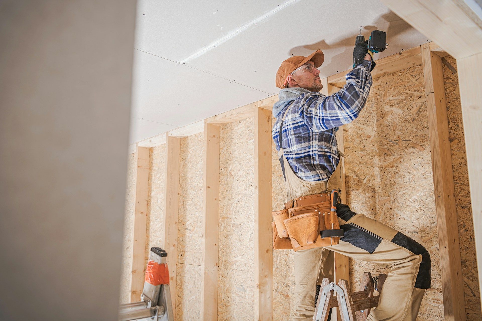 Worker installing plywood wall panels on a ladder in an unfinished room.