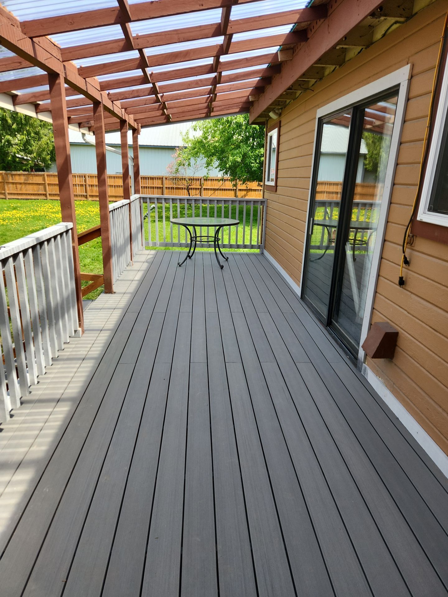 Wooden deck walkway beside a house, with railing, pergola beams, sliding glass door, and a patio table at the end