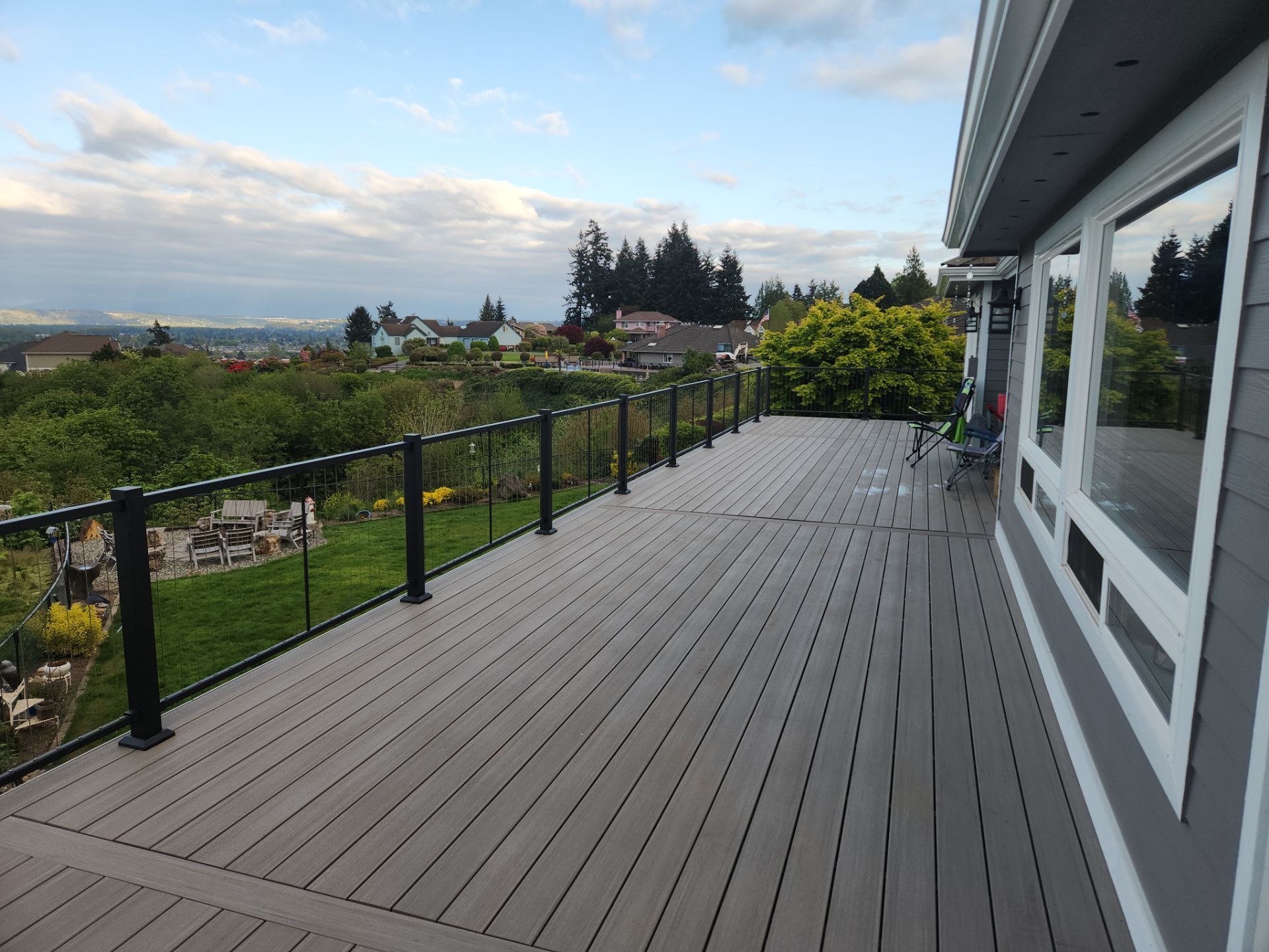 Wide gray deck with black railing beside a house, overlooking a green yard and cloudy sky.