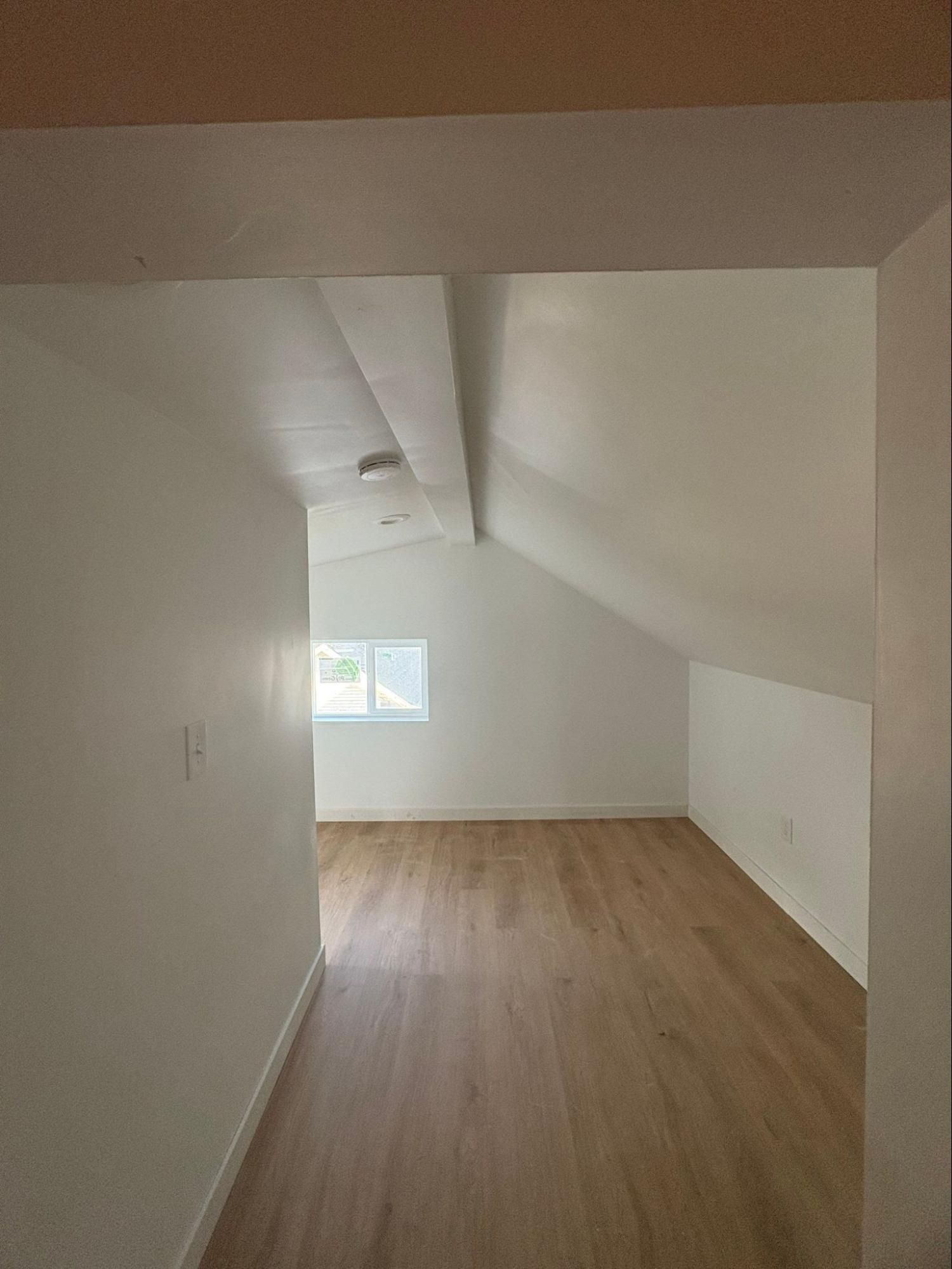 Empty narrow attic room with white walls, sloped ceiling, wood floor, and a small window at the far end