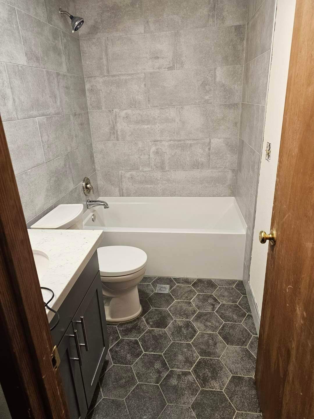 Small bathroom with gray tiled walls, white tub, toilet, dark vanity, and patterned floor.
