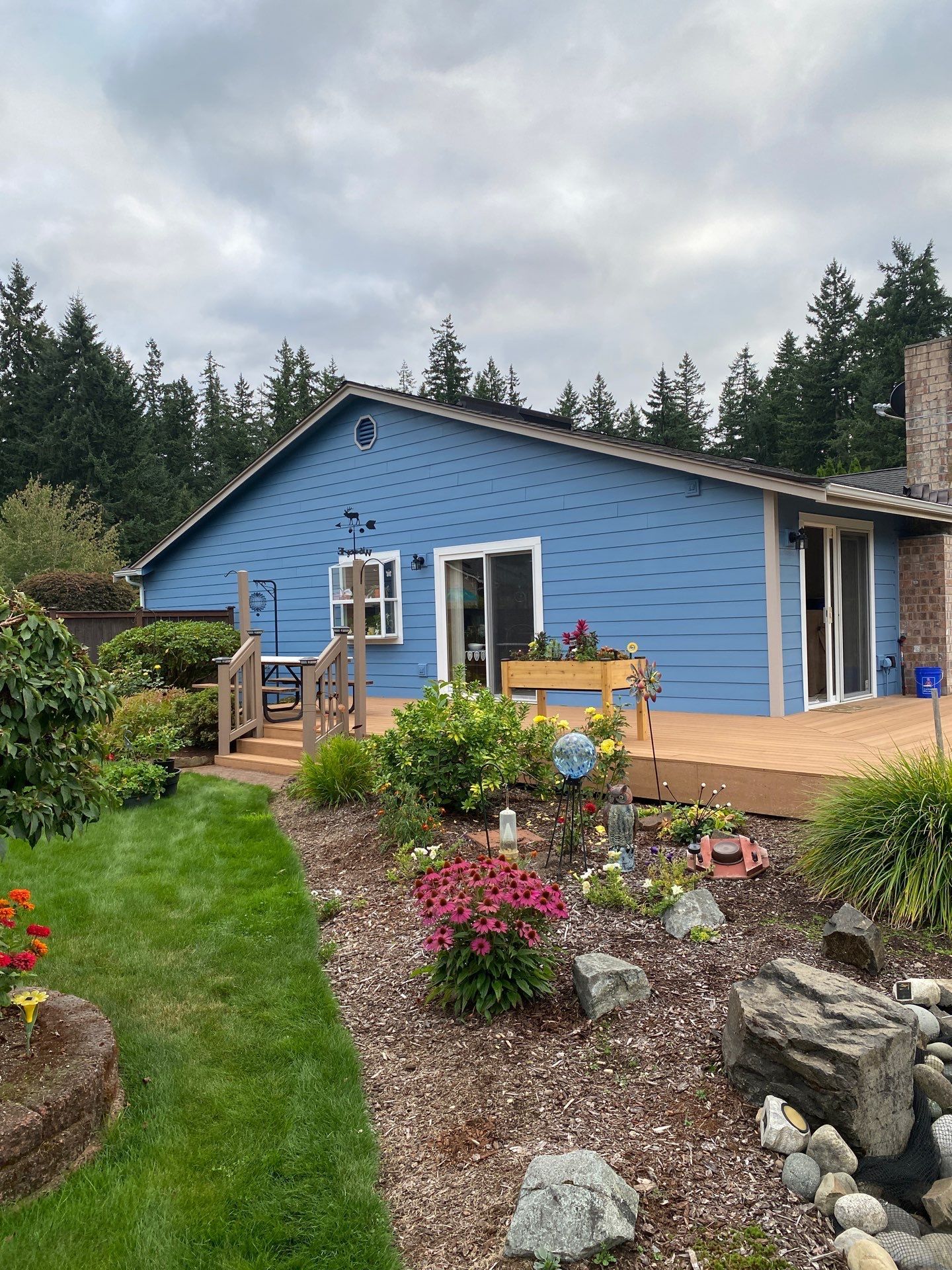 Blue house with deck and garden, surrounded by trees under a cloudy sky