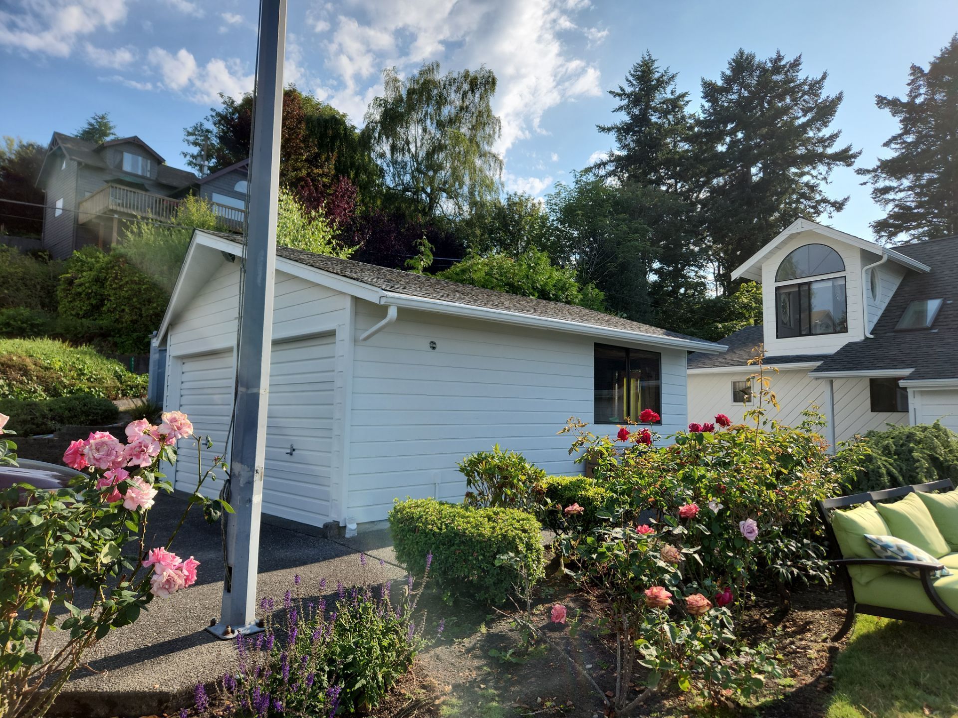 White garden shed with flowers and shrubs in a sunny backyard, with trees and a house in the background