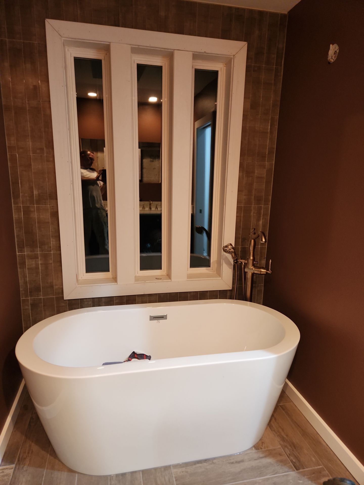 Freestanding white bathtub in a tiled bathroom with a triple-pane window above it