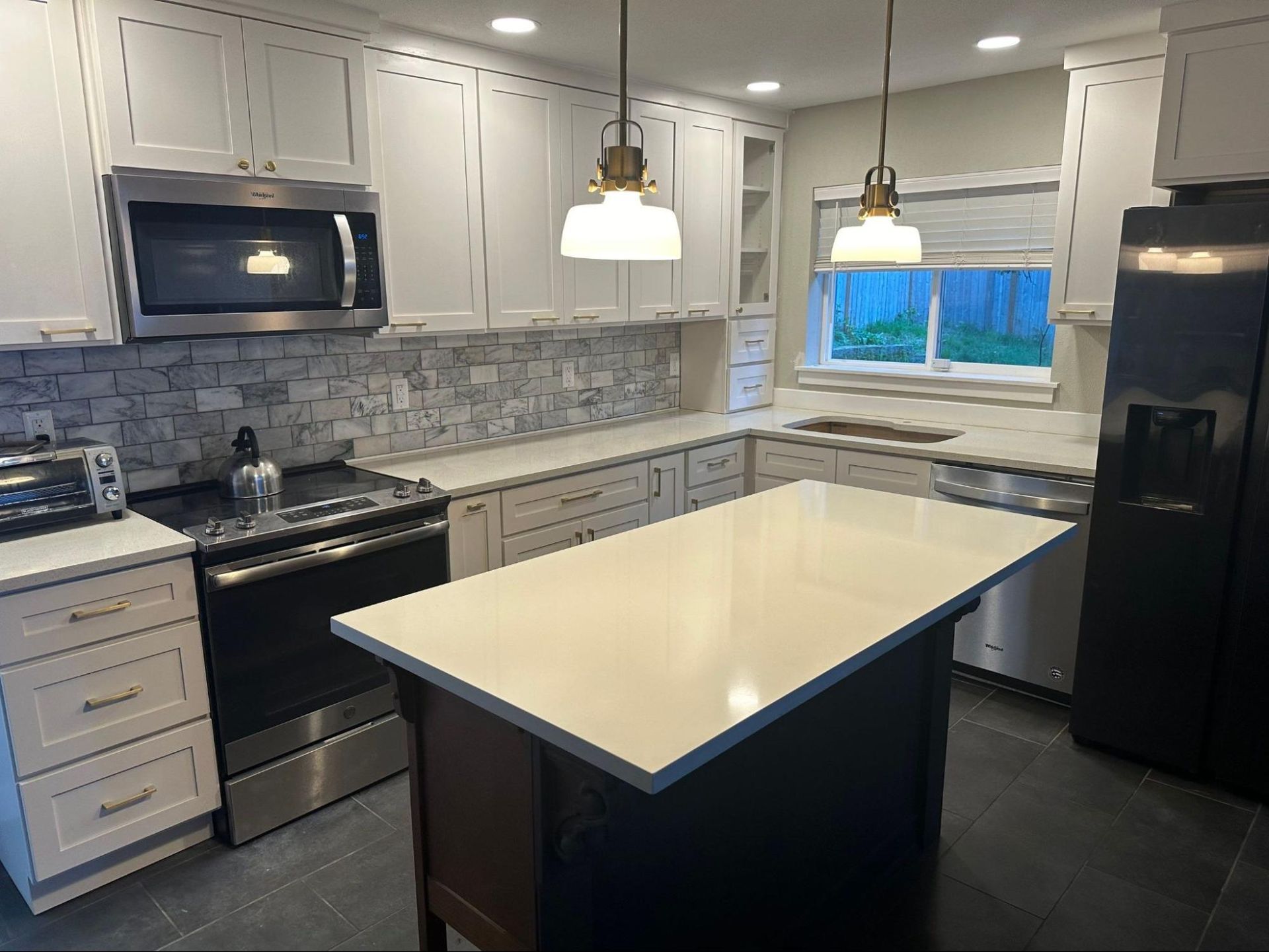Modern white kitchen with island, black appliances, gray tile backsplash, and a window over the sink.