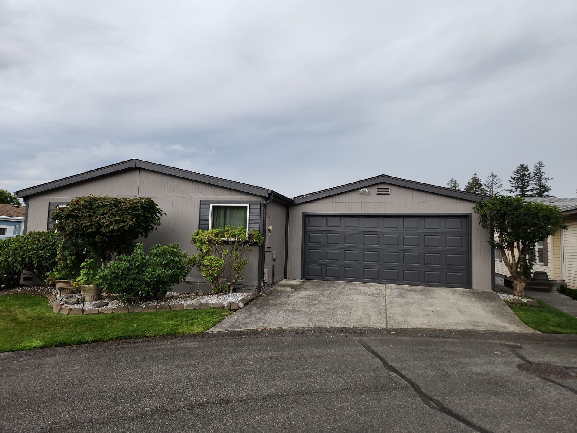 Single-story gray house with a dark garage, driveway, and shrubs under a cloudy sky