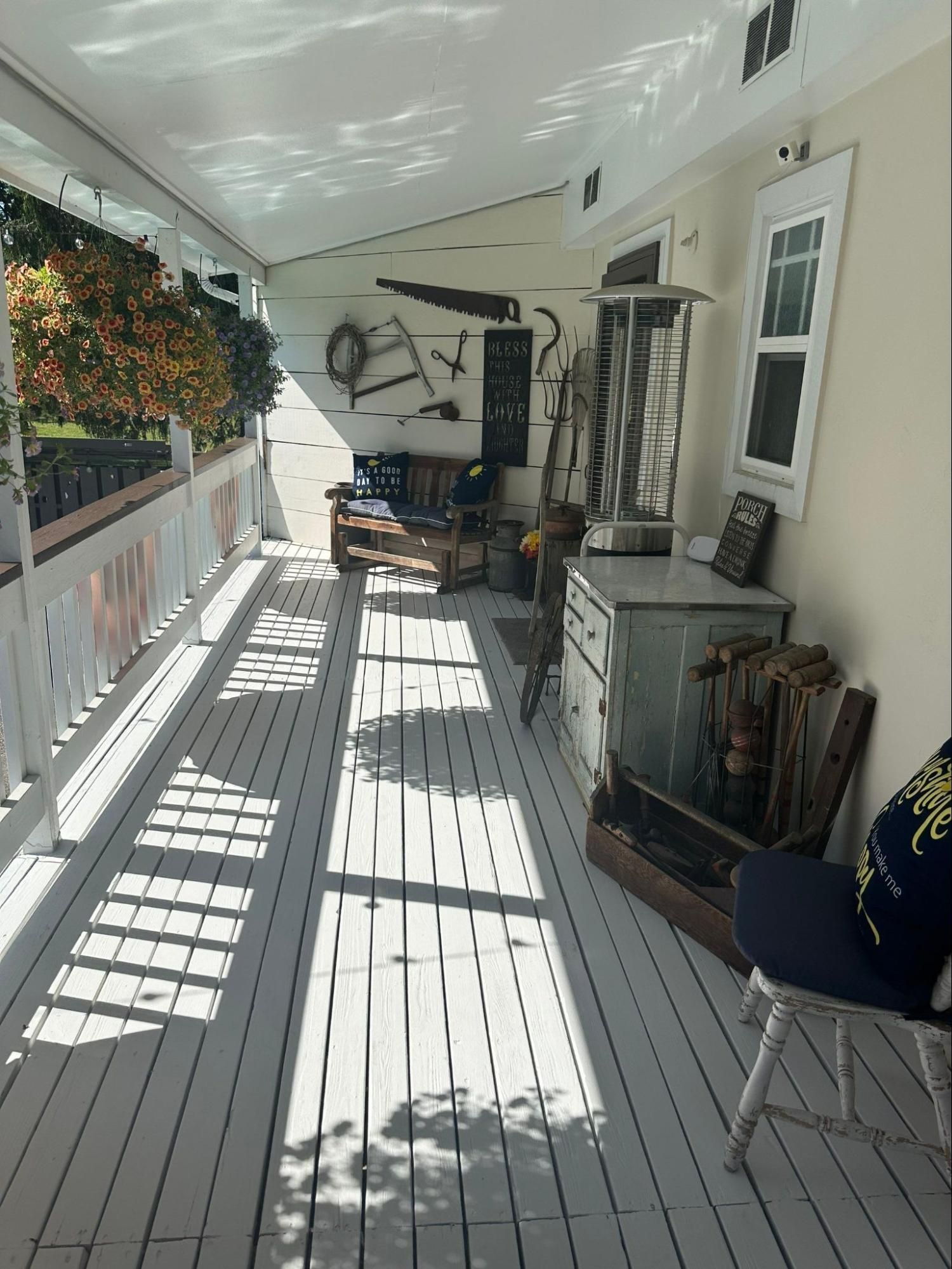 Sunlit porch with white railing, gray floor, hanging flowers, and chairs along the wall.
