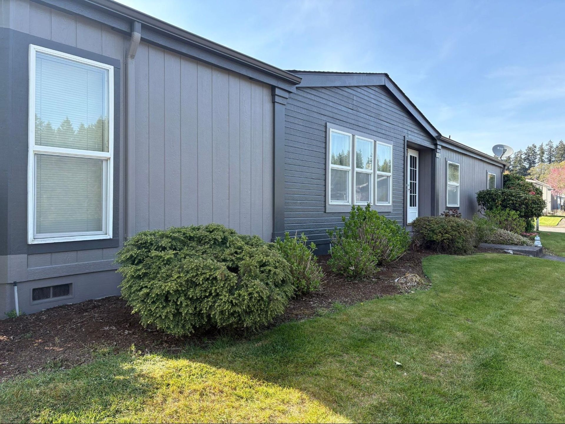 Gray house exterior with white-trimmed windows and shrubs along a green lawn