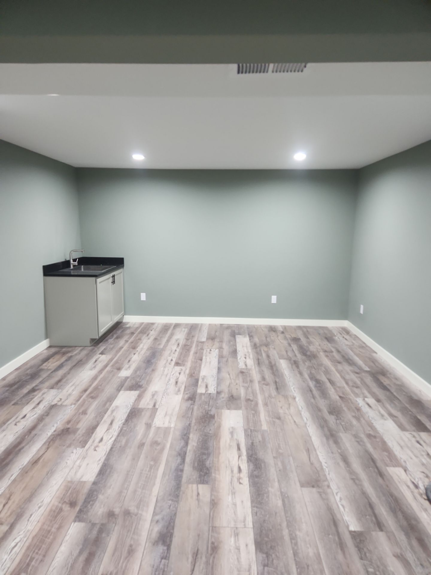 Empty finished basement room with gray walls, light wood floors, recessed ceiling lights, and a small kitchenette corner.