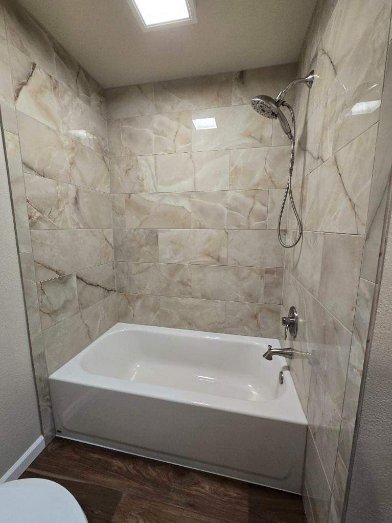 Small beige-tiled bathroom with a white tub, showerhead, and dark wood floor.